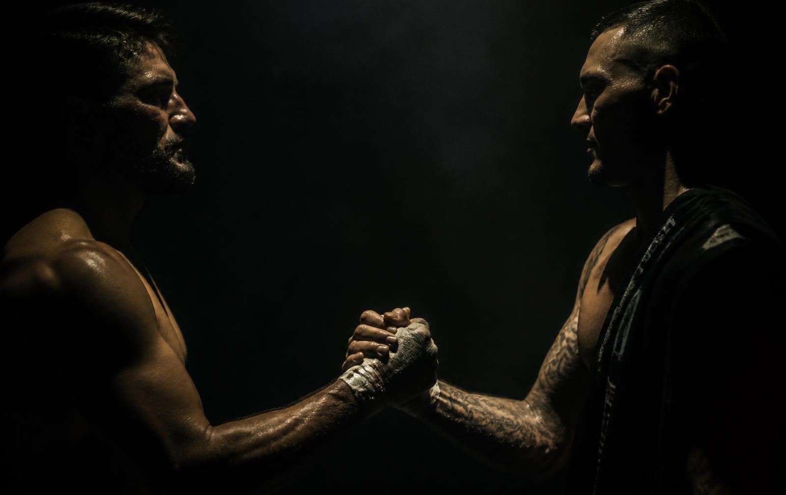 Two athletes clasping forearms in a warrior handshake, lit dramatically against darkness